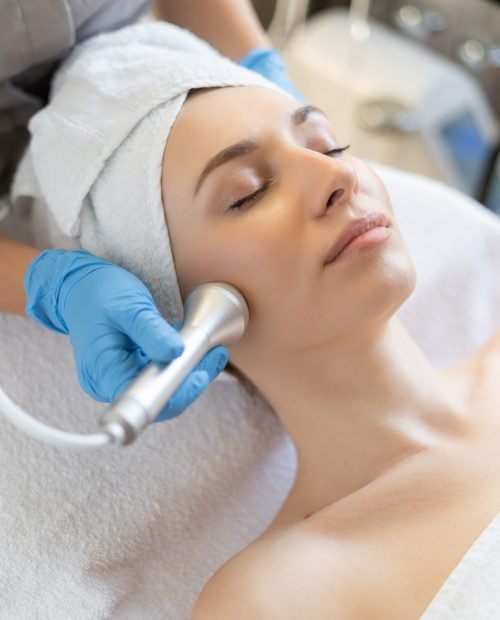 Young woman lying on cosmetologist's table during rejuvenation procedure. Cosmetologist take care about neck and face skin youthfull and wellness. Hardware face cleaning procedure